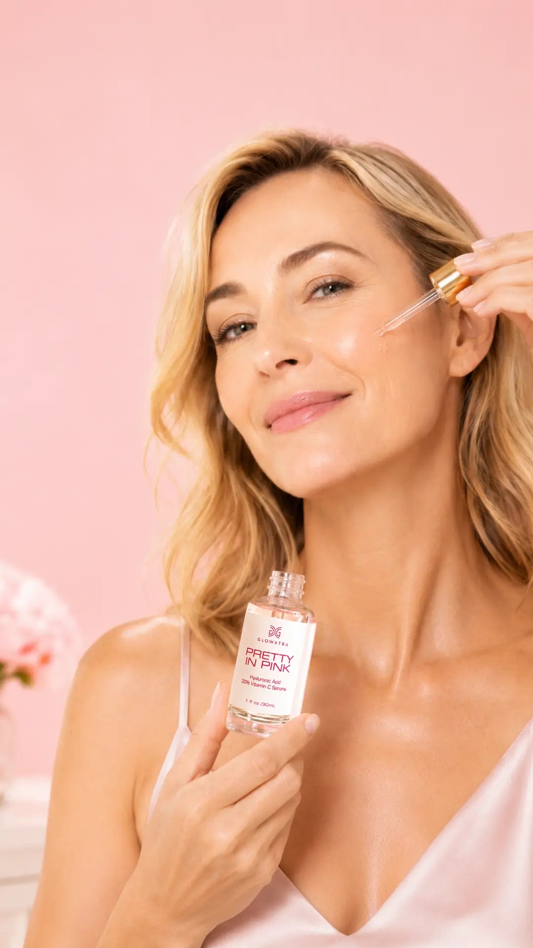 A woman with blonde hair applies a skincare serum dropper to her face while holding a clear bottle labeled 'Glowing Skin Serum' against a soft pink background.